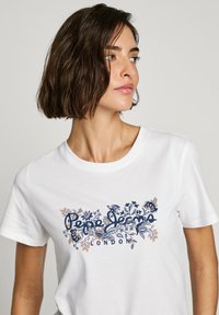 Vit bomull t-shirt med blommönster i marinblått och orange, med texten "Pepe Jeans" och "LONDON." Kort ärm, rundad nacke.