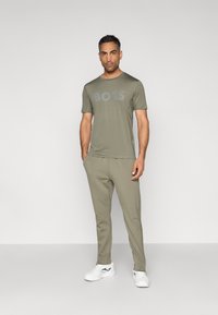 T-shirt verde-oliva de manga curta com um grande logótipo "BOSS" em cinza claro, combinada com calças de treino olive verde e ténis brancos.