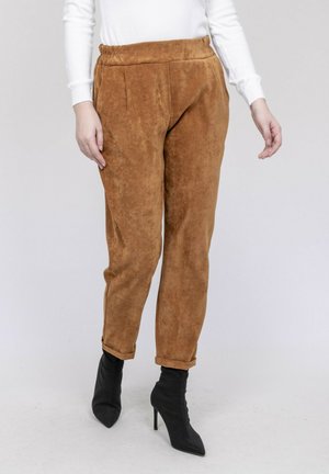 Personne portant un pantalon en daim beige et des bottes à talons aiguilles noires, debout devant un fond uni.