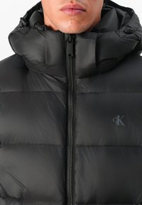 Zwarte gewatteerde vest met hoge kraag, voorzien van een ritssluiting aan de voorkant en een subtiel logo. Het materiaal heeft een gladde, glanzende textuur.