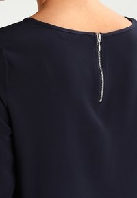 Top blu navy con scollatura rotonda e una cerniera metallica visibile sul retro. Il tessuto ha una consistenza liscia con dettagli sottili.