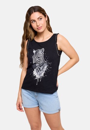 Joven con cabello largo y ondulado, lleva una camiseta negra sin mangas con un gráfico de cebra y pantalones cortos de mezclilla azul claro con dobladillo.