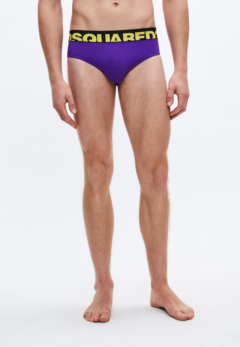 Lila férfi úszónadrág fekete derékszalaggal, melyen sárga "Dsquared2" logó található. Az anyag simának és rugalmasnak tűnik.