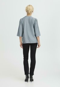 Helles graues Oberteil mit Dreiviertelärmeln, gerafften Schultern und geradem Schnitt. Kombiniert mit schwarzen, figurbetonten Jeans und Stiefeletten.