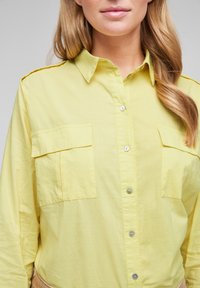Camicia gialla con colletto, dotata di due grandi tasche sul petto e realizzata in un tessuto leggero e leggermente strutturato.