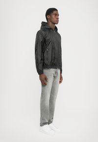 Svart zip-up jacka med justerbar huva, prydd med ett strukturerat mönster, tillsammans med grå jeans och vita sneakers.