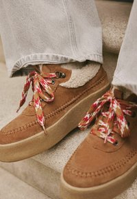 Brown suede platform shoes with patterned red, yellow, and brown laces. Sheepskin lining visible, showcasing a textured finish.