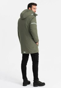 Parka verde con cappuccio e design a vita stretta, dotata di una striscia grigia sulle maniche, tasche laterali e un patch con logo in fondo. Pantaloni neri e stivali.