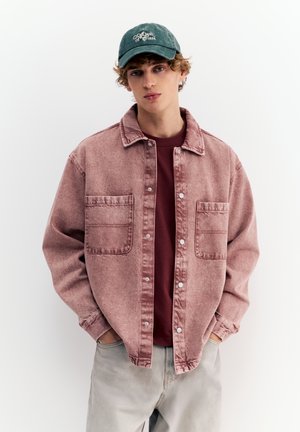 Mladý muž v zblednuto ružovej zapínanej bunde cez bordovú košeľu, svetlo sivých nohaviciach a tmavozelenej čiapke stojí s rukami v vreckách.