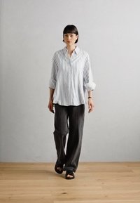 Camisa de botones a rayas azul claro y blanca con mangas remangadas, combinada con pantalones oscurecidos de corte holgado y sandalias negras.
