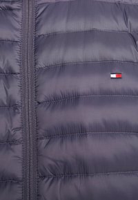 Giacca a vento grigia scura, con cerniera verticale, motivo trapuntato e un piccolo logo multicolore vicino alla cerniera.