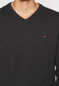 Maglione grigio scuro a scollo a V con una texture a maglia fine, dotato di un piccolo logo rosso, bianco e blu nell'area sinistra del petto.