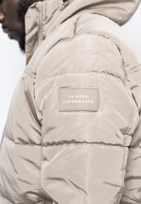 Giubbotto puffer beige con sezioni cucite, texture liscia e un'etichetta laterale con la scritta "LA ROSA COPENHAGEN" in testo bianco.