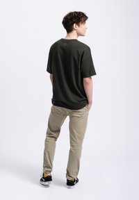 Donkergroene katoenen t-shirt met korte mouwen, relaxte pasvorm en een subtiel logo op de achterkant, gecombineerd met lichtbruine cargobroeken en zwarte sneakers.