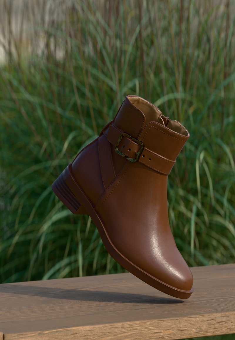 Bottine en cuir marron avec boucle latérale et petit talon, positionnée sur une surface en bois avec un fond d’herbe verte.