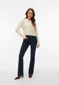 Krēmkrāsas kardigans ar apkakli, ar teksturētiem rotājumiem. Kombinēts ar tumši ziliem zvīļveida džinsiem un brūnām kurpēm ar papēžiem.