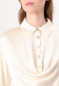 Blusa in satin crema con colletto classico, chiusura frontale con bottoni dorati e morbido drappeggio sullo scollo.