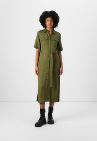 Abito camicia midi verde oliva con maniche corte, chiusura a bottoni, spacco laterale e cintura coordinata. Abbinato a stivali da combattimento neri con lacci.