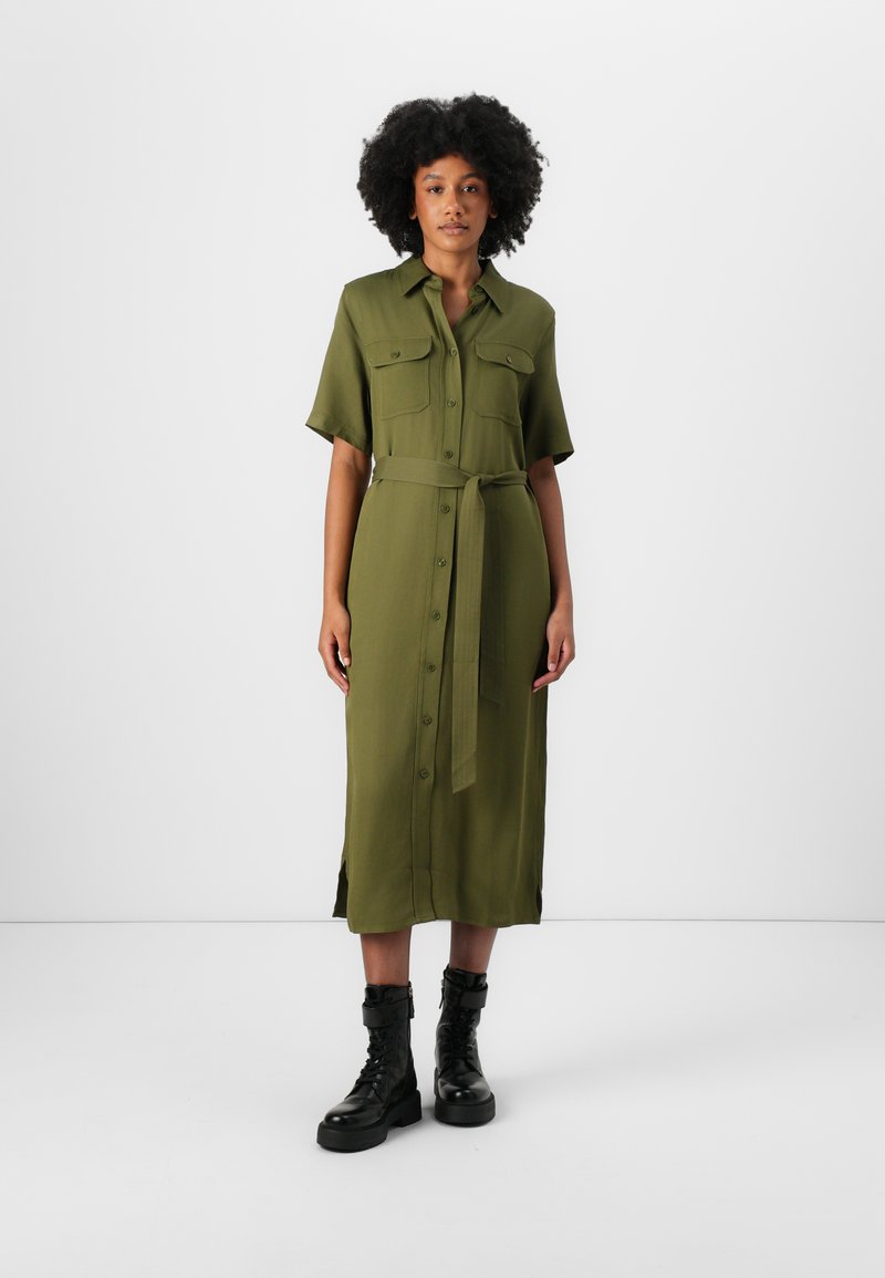 Abito camicia midi verde oliva con maniche corte, chiusura a bottoni, spacco laterale e cintura coordinata. Abbinato a stivali da combattimento neri con lacci.