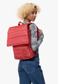 Mochila roja de forma cuadrada, material texturizado, cierre de solapa y un diseño gráfico con temática de cacahuete y logotipo en la parte frontal.