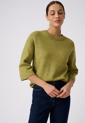 Vrouw met donker haar die een olijfgroene gebreide trui met driekwartmouwen en donkerblauwe jeans draagt, staand tegen een witte achtergrond.