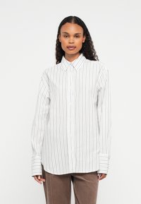 Vrouw met lang krullend haar, gekleed in een witte blouse met zwarte verticale strepen en bruine broek, staande tegen een effen witte achtergrond.