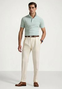 Camisa polo verde claro com gola, combinada com calças plissadas bege e sapatos de couro castanho. O conjunto inclui um cinto e um relógio castanhos.