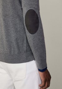 Pull en maille grise avec des patchs de coudes noirs contrastants, des poignets et un ourlet côtelés. Associé à un pantalon blanc avec une finition texturée.