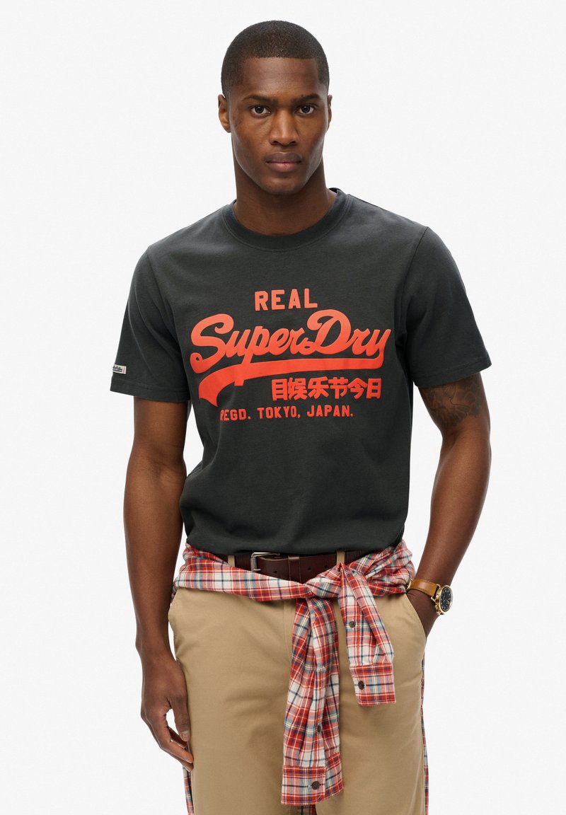 Camiseta de algodón negra con gráfico rojo de "SuperDry". Se lleva con pantalones beige y una camisa de cuadros rojos atada a la cintura. Mangas cortas.