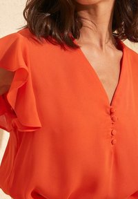 Blouse rouge avec manches à volants, décolleté en V et boutons décoratifs sur le devant. Confectionnée dans un tissu fluide, offrant une texture douce.