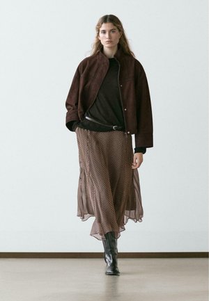 Donna che cammina al chiuso indossando una giacca marrone scuro, top nero, gonna marrone con motivo, stivali neri, contro uno sfondo chiaro e uniforme.