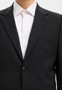 Czarna marynarka z wyciętymi klapami, zapinana na jeden guzik, z kieszenią na piersi, noszona na białej koszuli wizytowej z widocznymi guzikami.