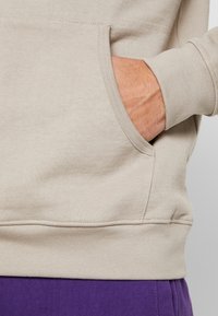 Ruka odpočívaná uvnitř přední kapsy béžového svetru, kombinovaného s fialovými kalhotami, ukazuje texturu látky a detaily švů.