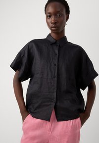 Camicia nera a maniche corte con bottoni, realizzata in un tessuto leggero e testurizzato, con colletto e orlo dritto. Abbinata a pantaloni rosa con motivo.