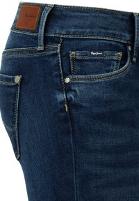 Σκούρο μπλε τζιν με ορατές μπροστινές και πλαϊνές τσέπες, κίτρινα ραφή, μεταλλικά καρφιά και καφέ δερμάτινη ετικέτα επωνυμίας στη μέση.