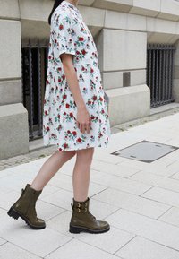 Abito floreale bianco con rose rosse e foglie verdi, maniche corte, abbinato a stivaletti alla caviglia verde oliva con lacci e fibbie dorate.