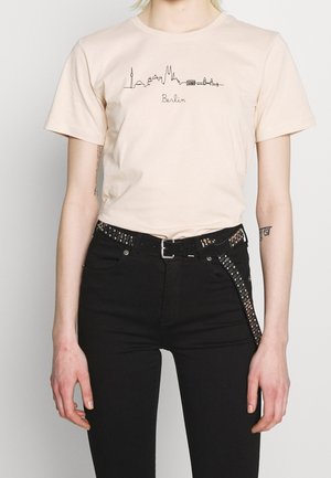 Osoba nosząca jasnobeżową koszulkę z czarną panoramą Berlina i tekstem, w połączeniu z czarnymi spodniami i paskiem z ćwiekami jako dodatkiem.