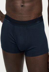 Άνδρας με σκούρο δέρμα, φορώντας ναυτικό μπλε μποξεράκι Calvin Klein με λογότυπο στη μέση, στέκεται μπροστά σε ουδέτερο φόντο.