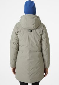Lichtgrijze geïsoleerde jas met een capuchon, voorzien van een zwart logo op de achterkant, een gladde textuur en een lange lengte, gecombineerd met een blauwe gebreide beanie.