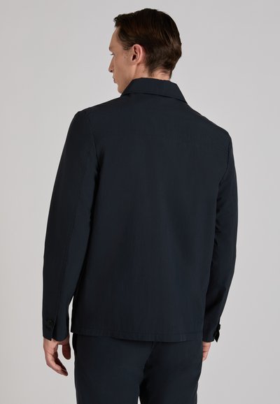 Homme vu de dos portant une veste à manches longues bleu marine foncé et un pantalon assorti, sur un fond gris clair uni.
