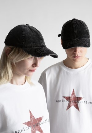 Dwie osoby w czarnych czapkach i białych koszulkach z dużą czerwoną gwiazdą i napisem „against perfection” na przodzie, patrzące lekko w dół.