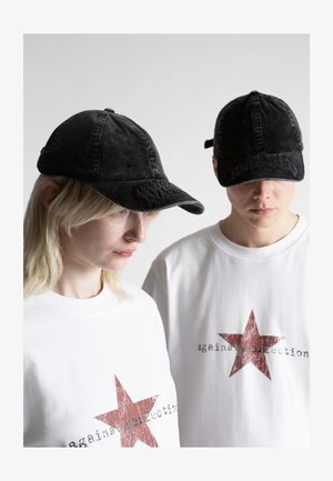 Du žmonės, dėvintys juodus kepurėlius ir baltus marškinėlius, ant kurių priekyje yra didelė raudona žvaigždė ir užrašas „against perfection“, žvelgia truputį žemyn.