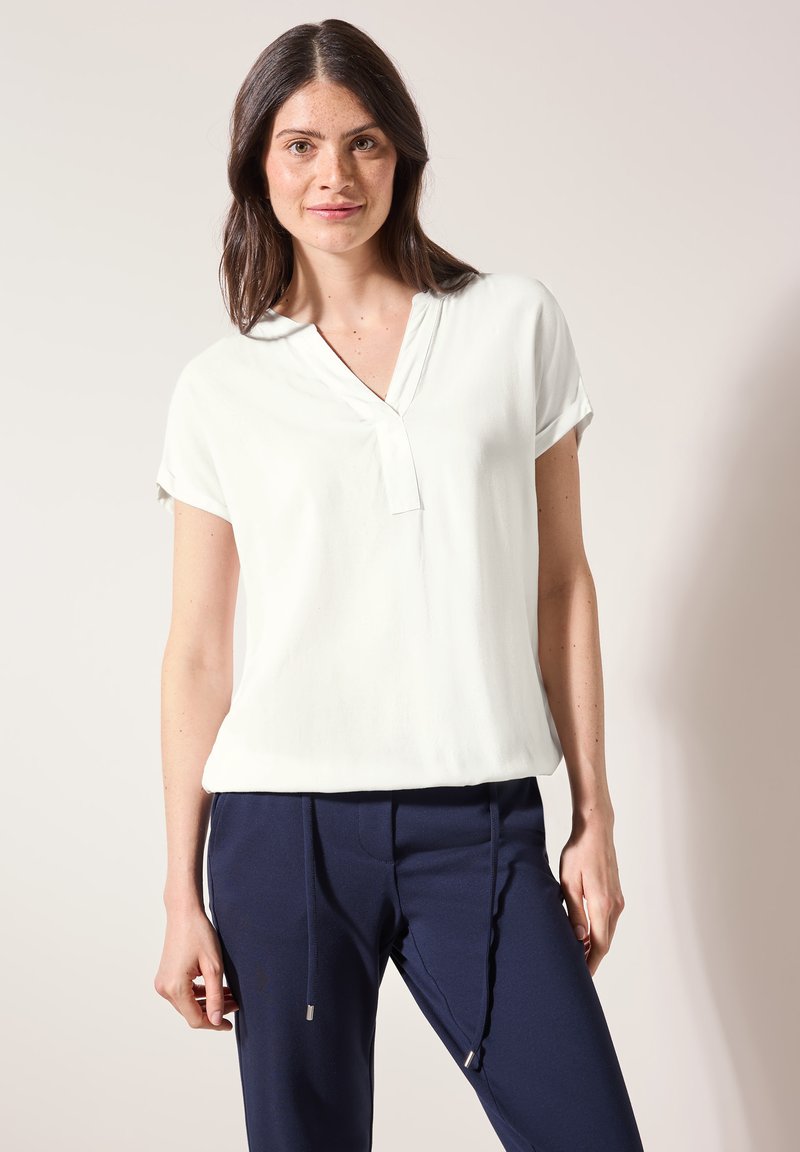 Blouse blanche à manches courtes avec un col en V et une coupe décontractée. Le tissu semble lisse et léger, complété par un pantalon bleu marine avec cordons de serrage.