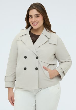 Hymyilevä nainen, jolla on vaaleanruskeat hiukset, pukeutunut beigeen kaksiriviseen takkiin ja valkoisiin housuihin, oikea käsi taskussa.