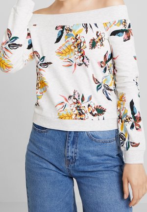 Femme portant un pull blanc à épaules dénudées avec un imprimé floral coloré, associé à un jean bleu taille haute.