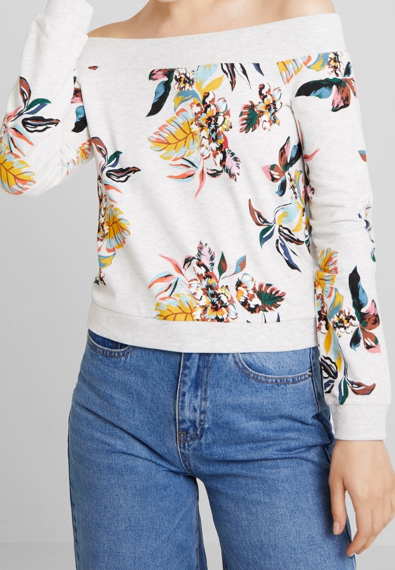 Femme portant un pull blanc à épaules dénudées avec un imprimé floral coloré, associé à un jean bleu taille haute.