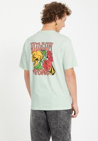 Camiseta verde menta con un gráfico colorido en la parte posterior que incluye una calavera, flores y la palabra "VOLCOM". Corte relajado, mangas cortas, material de algodón.