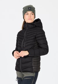 Sort pufferjakke med lynlås foran, der har horisontale quiltede sektioner og hætte. Sættes sammen med en grøn strikket beanie.