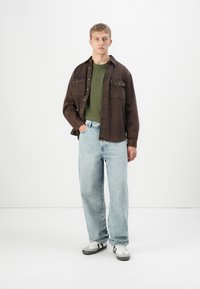 Young man stands against white background wearing brown overshirt, green ribbed sweater, light blue jeans, and white sneakers with black stripes.