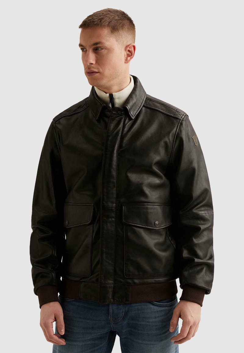 Mann trägt schwarze Lederjacke mit Klappentaschen und gerippten Bündchen, blickt nach links vor einfarbigem Hintergrund.